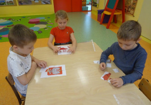 Troje dzieci siedzi przy stoliku i składa obrazek z części przedstawiający godło Polski.